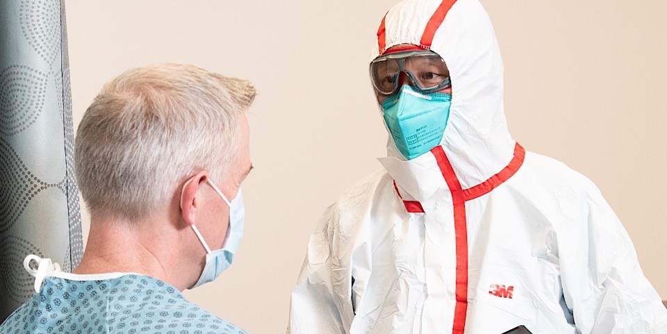 health care provider wearing full body suit, a particulate respirator and eye goggles talking with patient in mask
